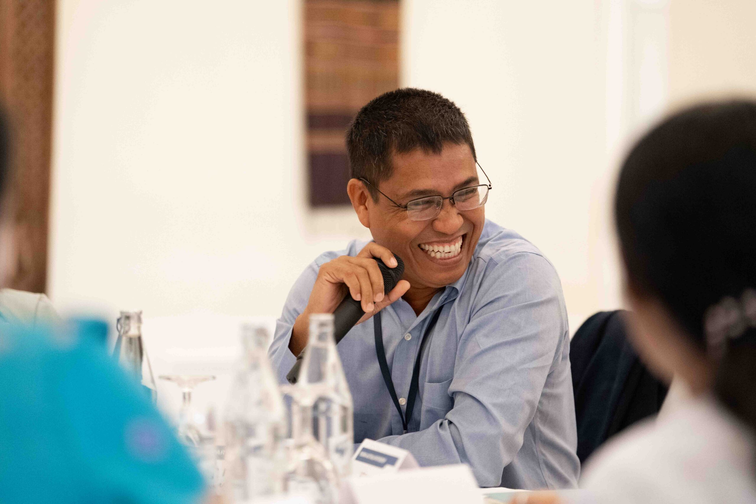 Photograph of a man smiling and holding a microphone during a discussion or meeting. Clear focus on the man wearing glasses and a light blue shirt, with blurred participants and water bottles in foreground and background.