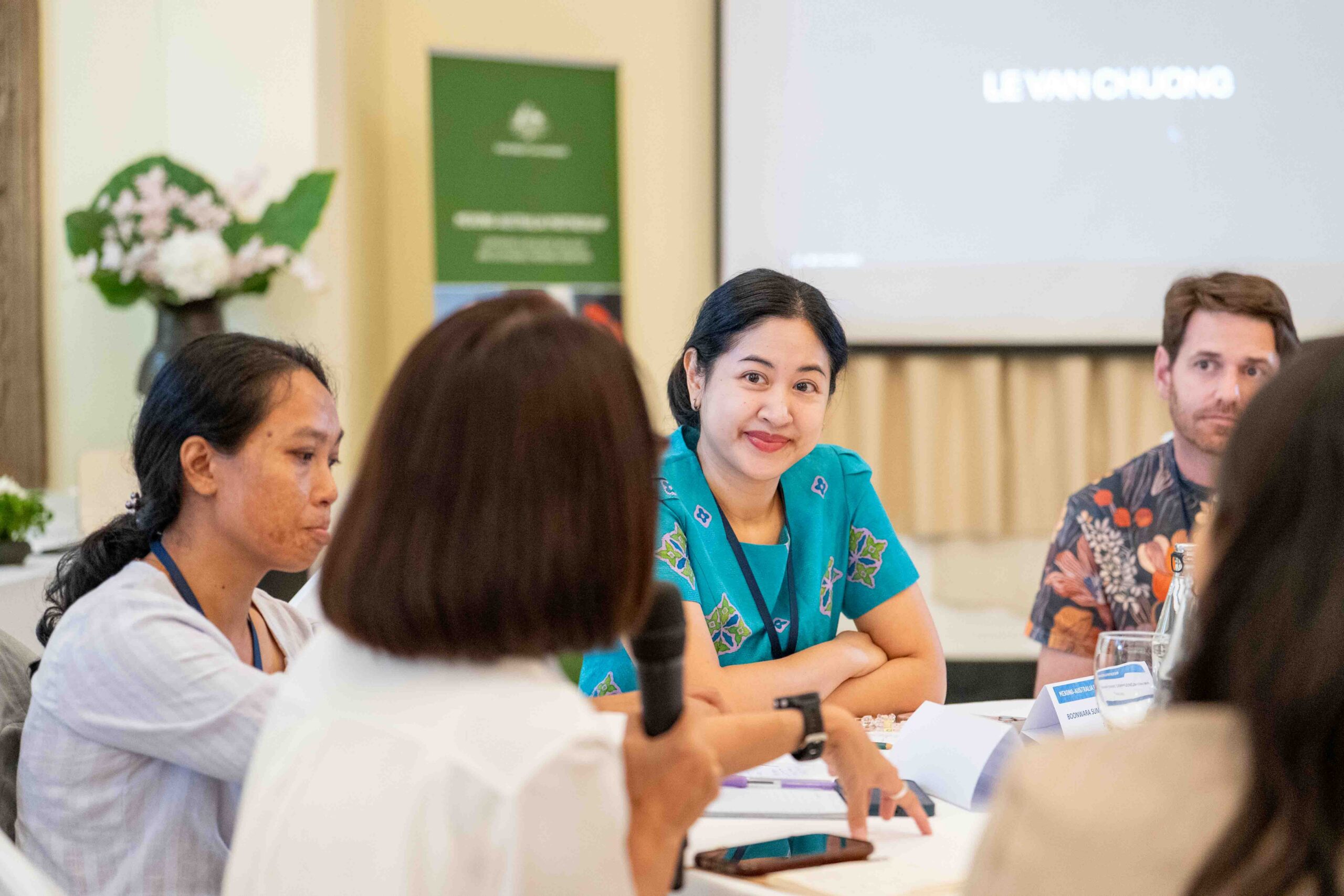 Photograph of a group discussion or meeting with four people seated around a table, one holding a microphone. Focus is on a woman in a teal shirt smiling, with name tags and a presentation screen in the background, suggesting a formal or professional setting.