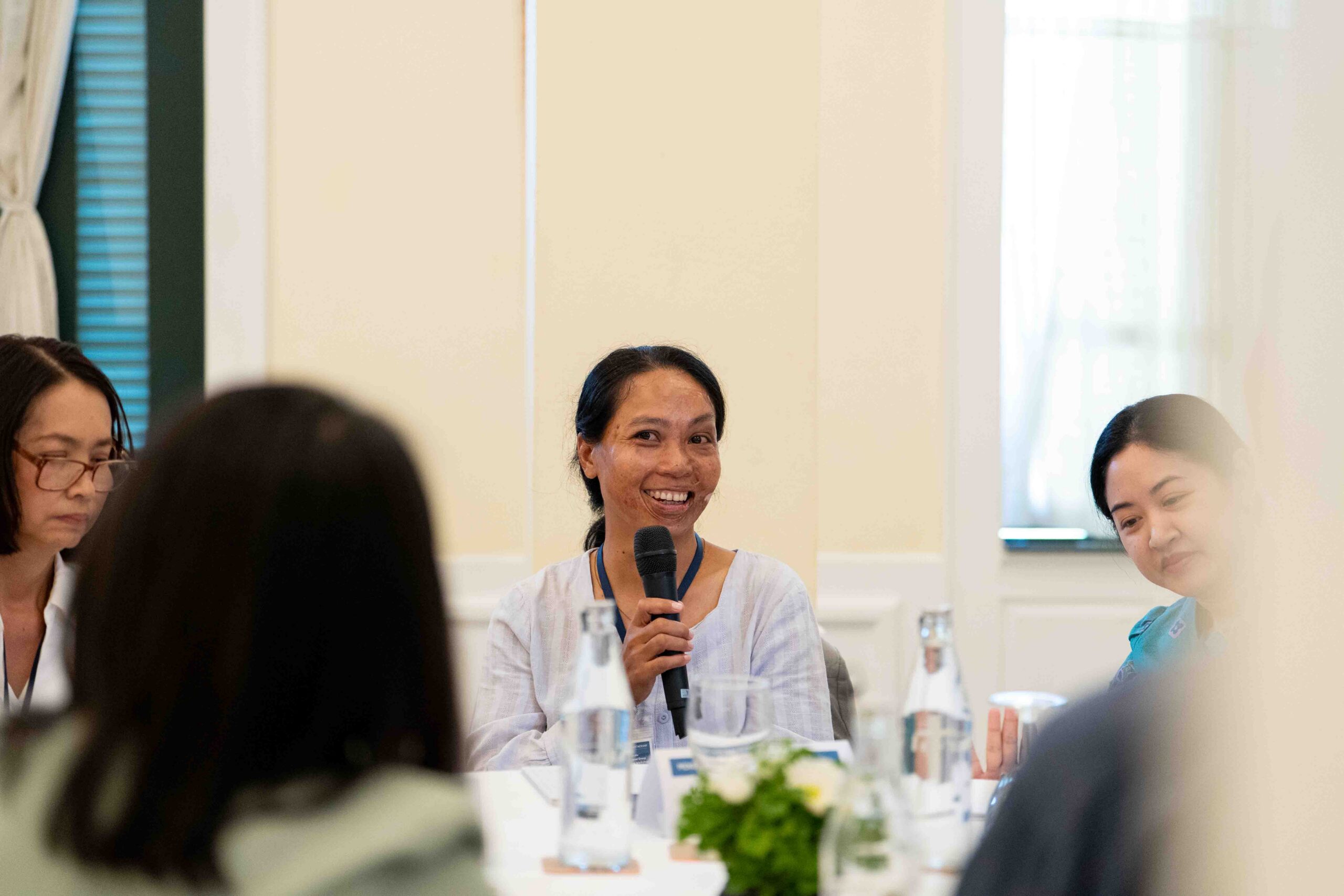 Photograph of a woman speaking into a microphone during a meeting or discussion, with three other people partially visible around a table. The setting includes water bottles, a small floral centerpiece, and natural light from a window, suggesting a formal yet relaxed group conversation.