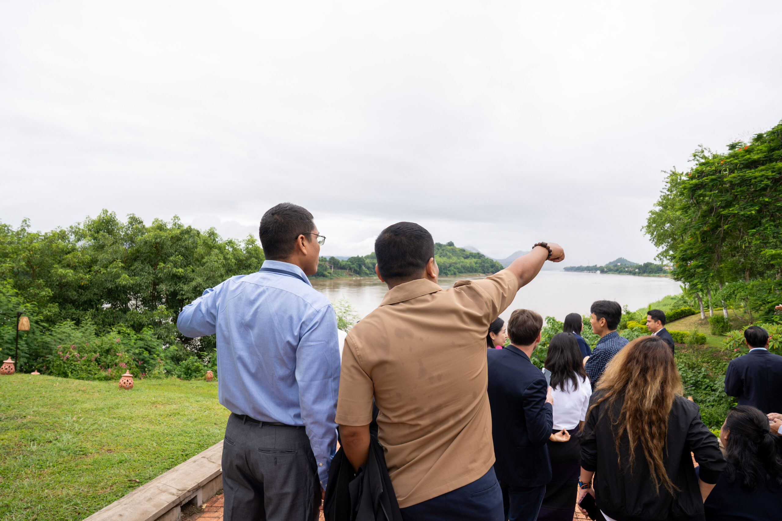Photograph of a group of people outdoors near a body of water, with two men in the foreground pointing towards the distance. The scene includes greenery, overcast sky, and individuals dressed in business casual attire, suggesting a professional or educational outing.