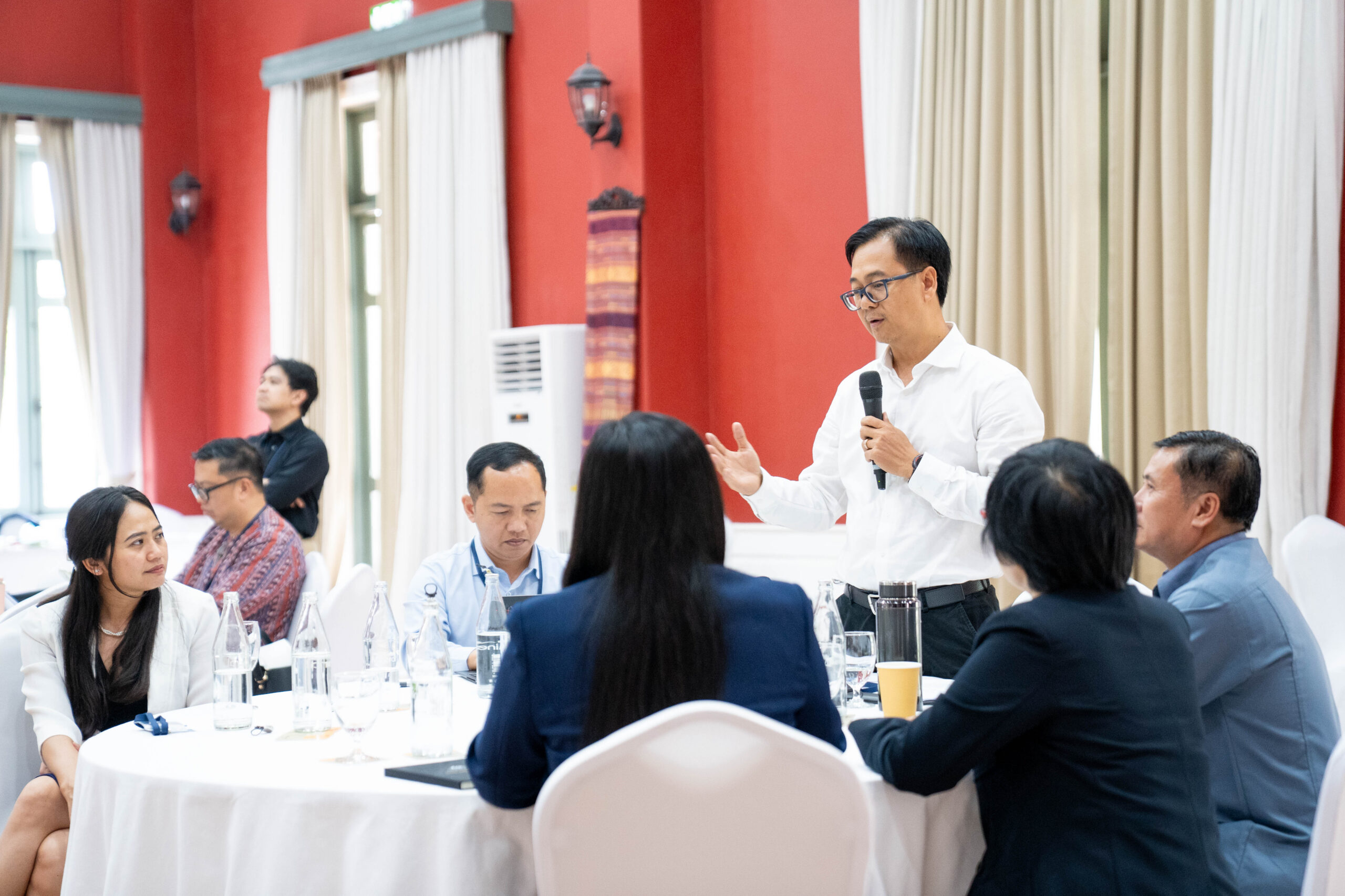 Photograph of a business meeting or conference with a man in a white shirt standing and speaking into a microphone while others seated around a round table listen attentively. The setting features red walls, white curtains, and formal attire, indicating a professional discussion or presentation.