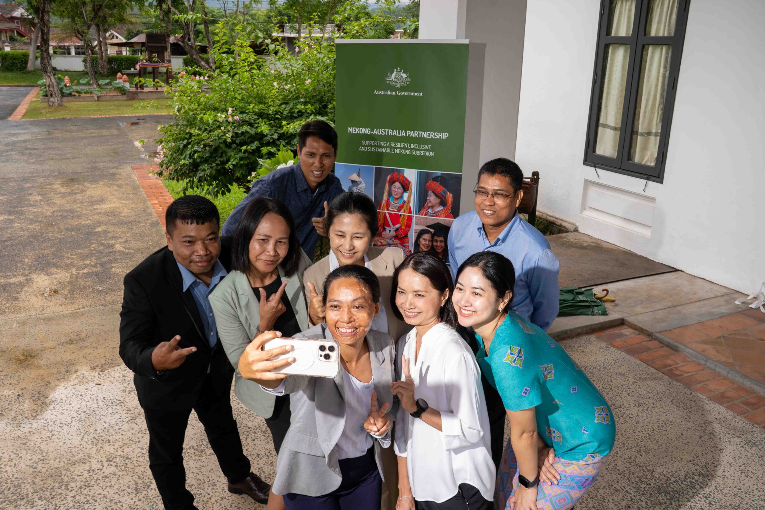 Photograph of a group of eight people outdoors taking a selfie in front of a building with a green banner displaying text and images related to a cultural or community event. Group includes men and women dressed in business casual and traditional attire, smiling and making peace signs, with greenery and a pathway visible in background.