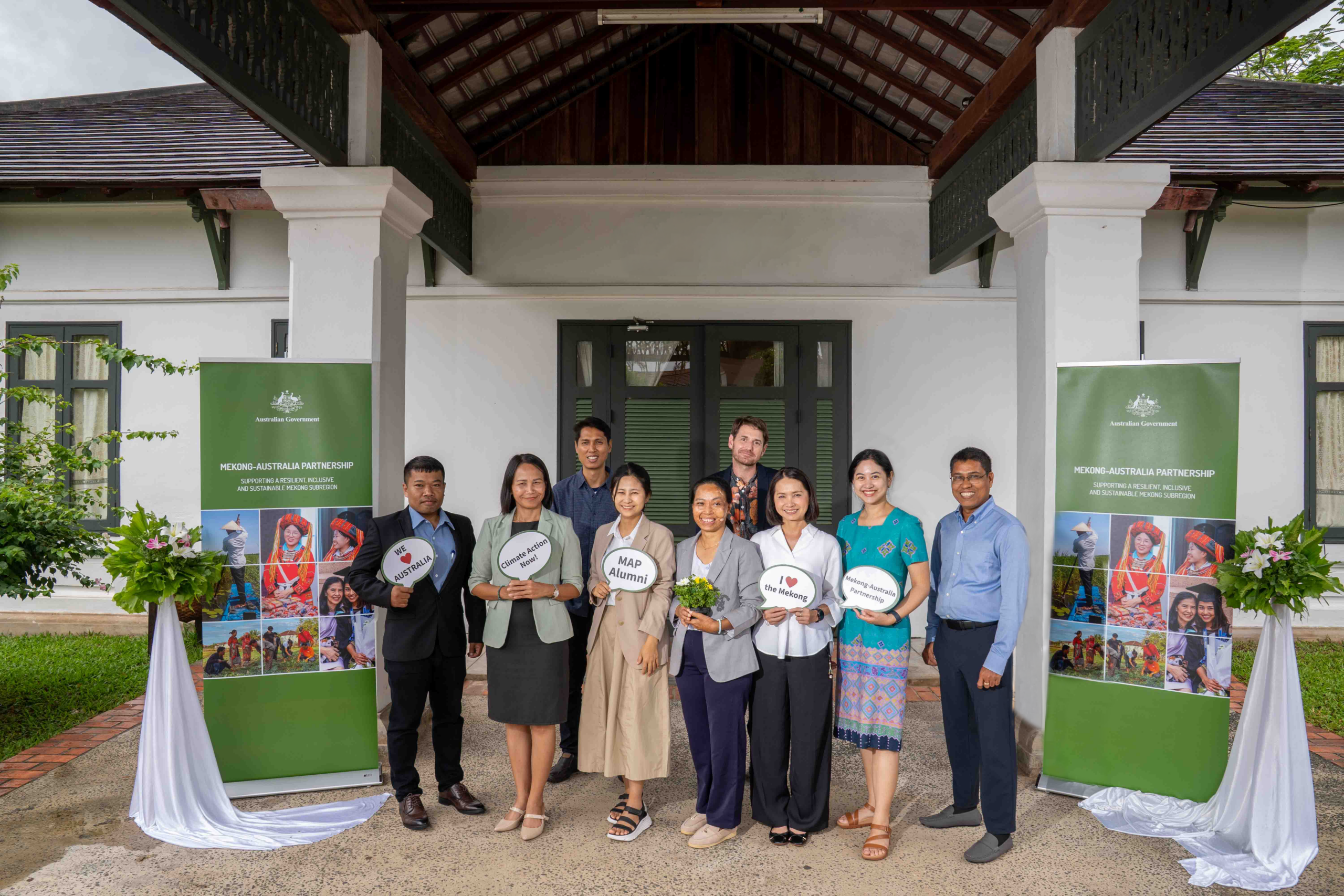 Photograph of a group of nine people standing in front of a building entrance with two green banners displaying cultural images and text about a program or event. Individuals hold signs with messages, and two floral arrangements with white drapes flank the group, indicating a formal or celebratory gathering.