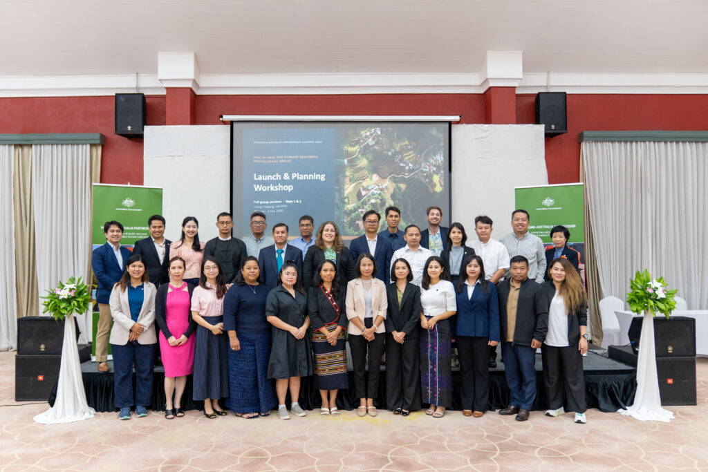 Photograph of a group of 25 people posing in two rows inside a conference room during a "Launch & Planning Workshop" event. The setting includes a projector screen displaying the event title, two green banners with logos on either side, and floral arrangements on white stands.