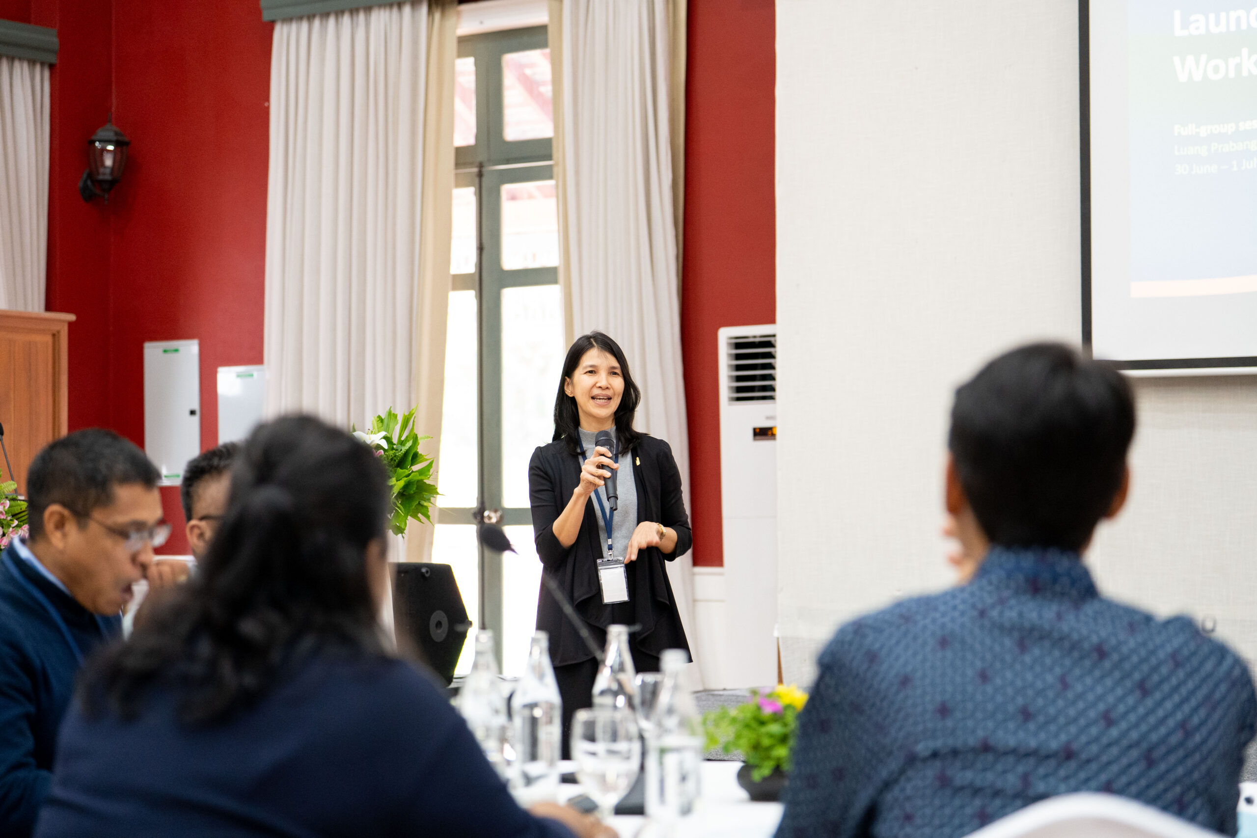 Photograph of a woman speaking into a microphone during a presentation in a conference room with red walls and large windows. Audience members are seated at a table in the foreground, with a projection screen displaying a slide in the background.