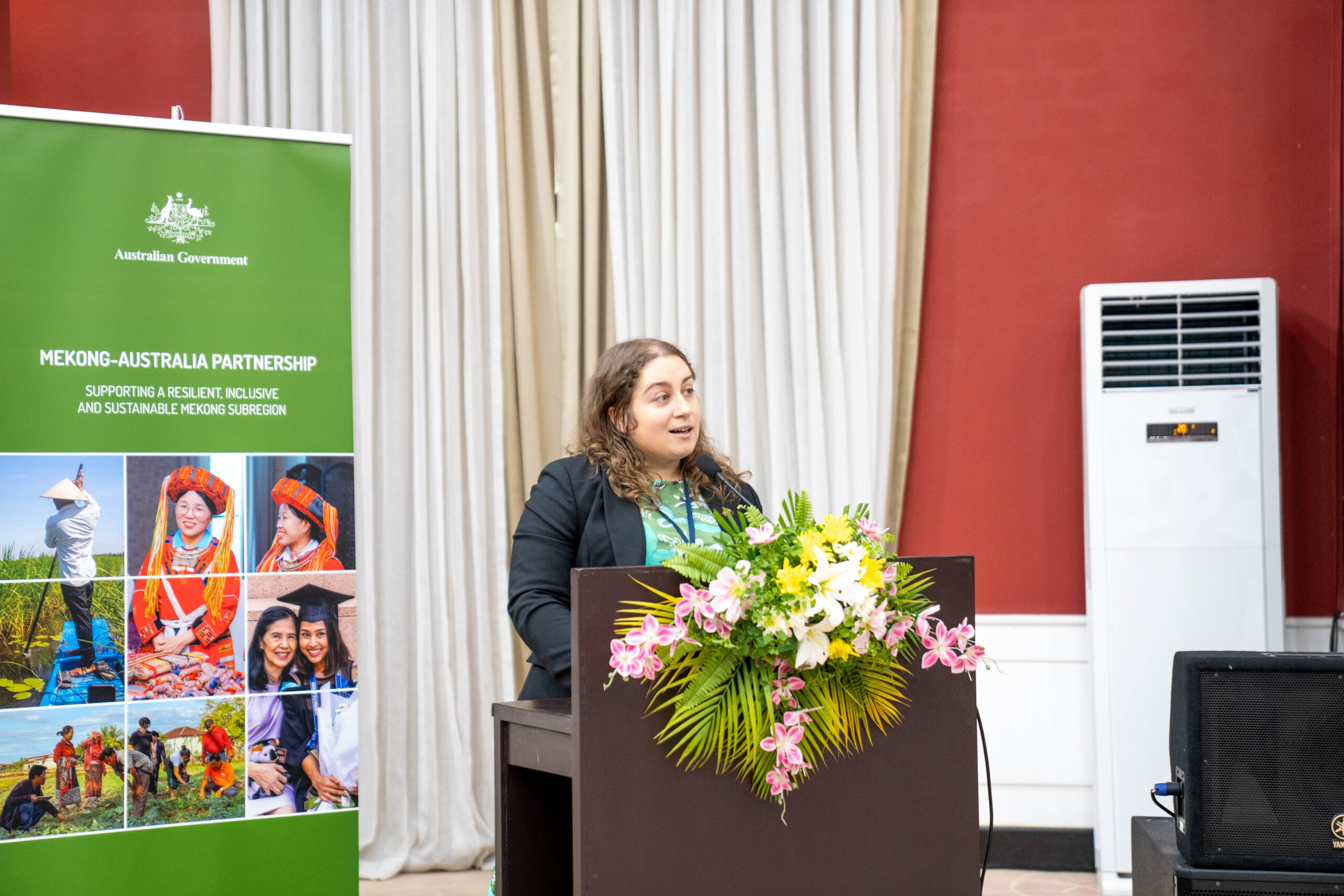 Photograph of a woman speaking at a podium decorated with a floral arrangement during an event. A green banner beside her displays "Mekong-Australia Partnership" with images of people and landscapes, indicating a focus on collaboration between Mekong region and Australia.