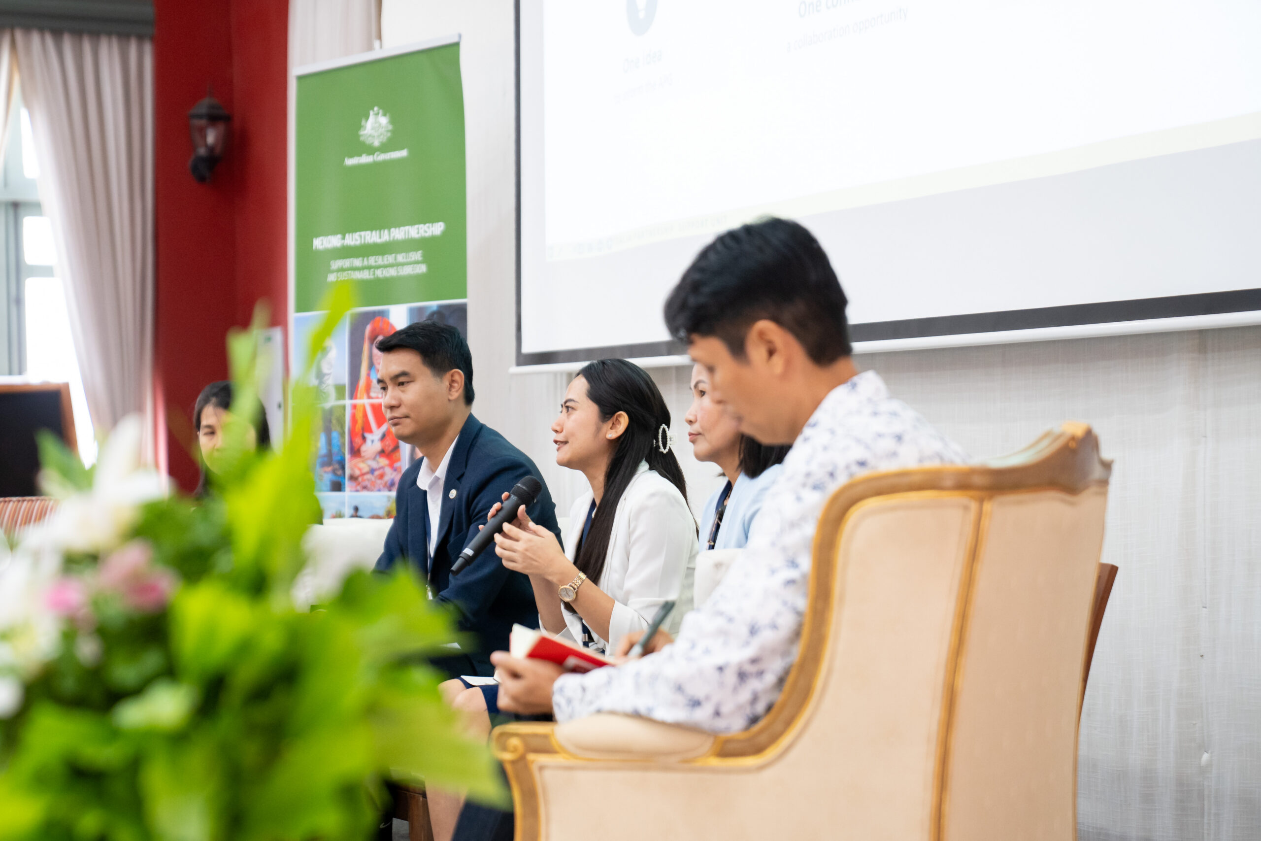 Photograph of a panel discussion featuring four individuals seated on stage, with one woman speaking into a microphone. Background includes a green banner and a white projection screen, while foreground shows blurred floral arrangements.